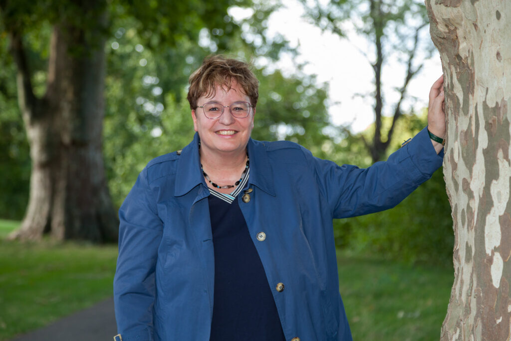 Rose Schliessmann – Coaching, Resilienztraining, Moderation und Naturangebote Dr. Rose Schließmann steht lächelnd neben einem Baum in grüner Natur und blickt in die Kamera.