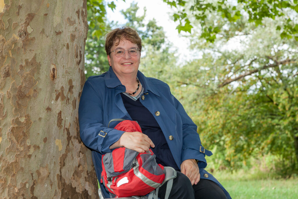 Rose Schliessmann – Waldbaden Dr. Rose Schließmann sitzt entspannt an einem Baum in grüner Natur.
