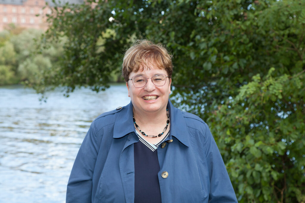 Dr. Rose Schließmann steht lächelnd am Ufer des Mains vor grünen Bäumen.